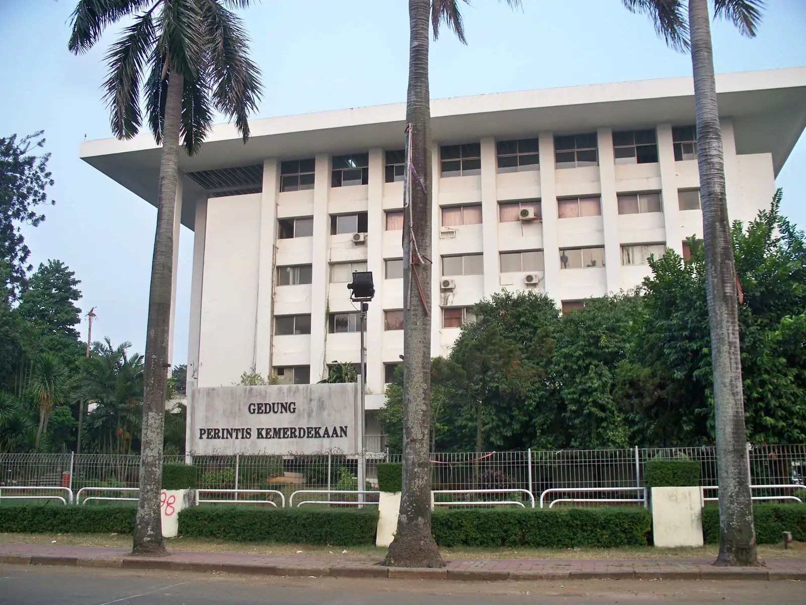 gedung perkantoran putih bertingkat dengan papan nama Gedung Perintis Kemerdekaan di balik pohon palem