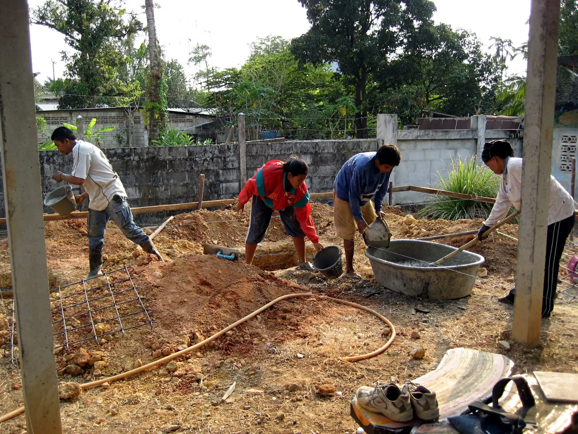 Empat pekerja mengaduk beton secara manual di bak adukan dan mengangkutnya dengan ember di proyek rumah