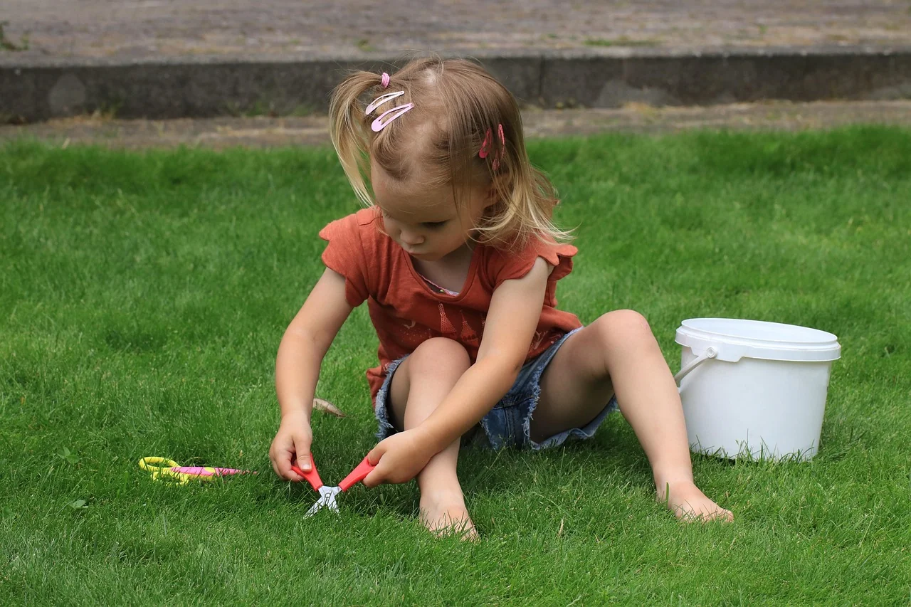 Anak kecil duduk bermain di atas rumput halaman dengan ember plastik dan gunting mainan