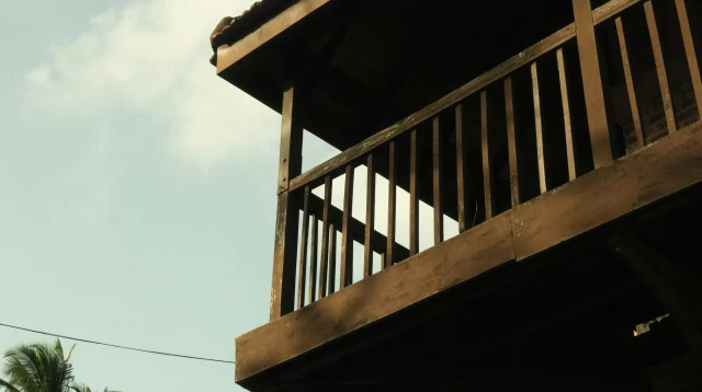 Lantai atas rumah dari kayu dengan balkon pagar kayu di antara pohon kelapa