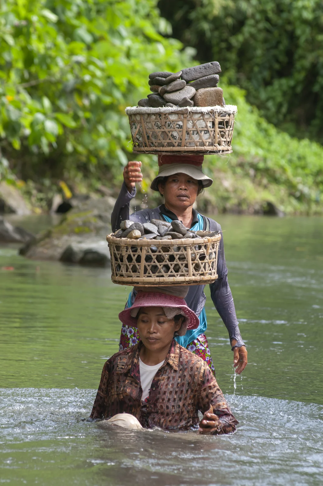 dua perempuan mengangkut batu kali dalam keranjang di atas kepala saat menyeberangi sungai