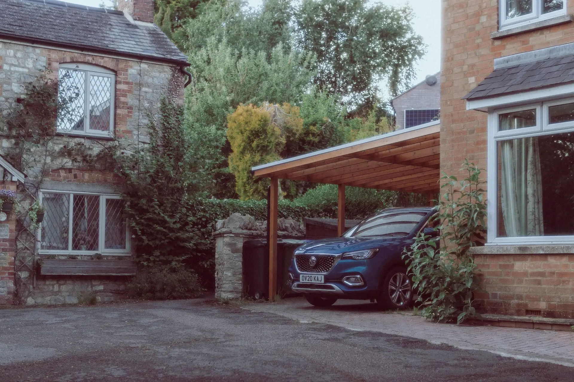 Carport kayu beratap miring di samping rumah bata dengan mobil SUV biru terparkir