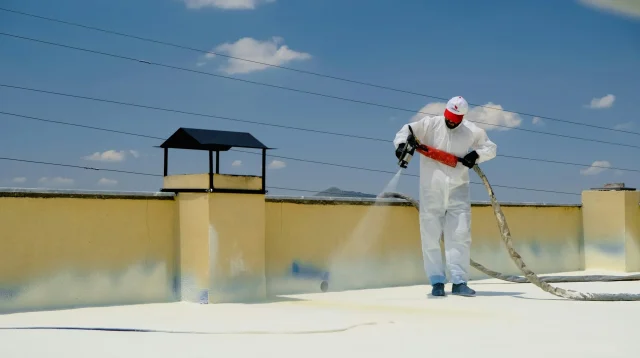 Pekerja berbaju pelindung menyemprotkan coating waterproofing putih di dak beton atap datar