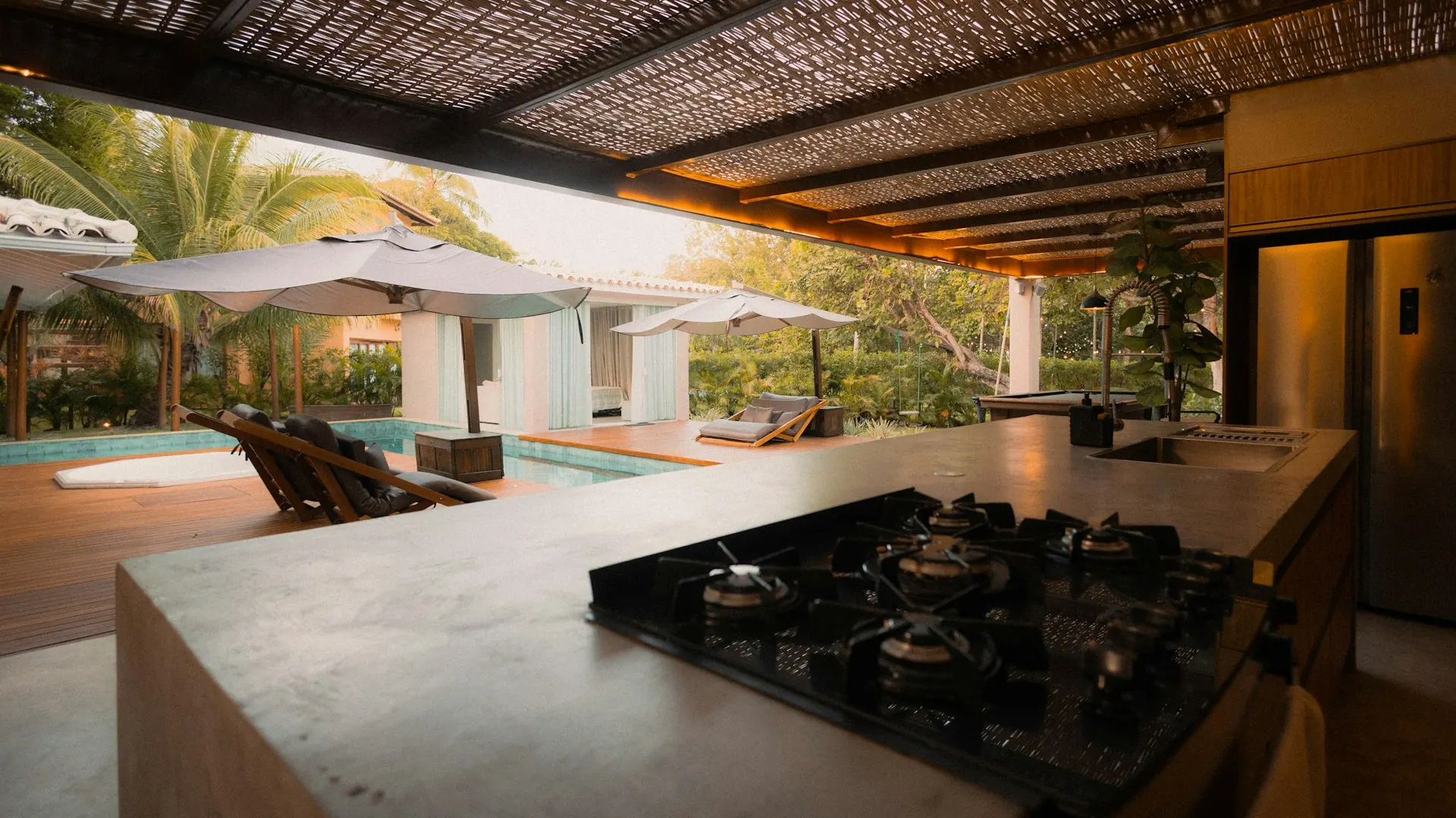 Dapur semi-terbuka di bawah pergola dengan kompor tanam, bak cuci, dan kulkas menghadap kolam renang