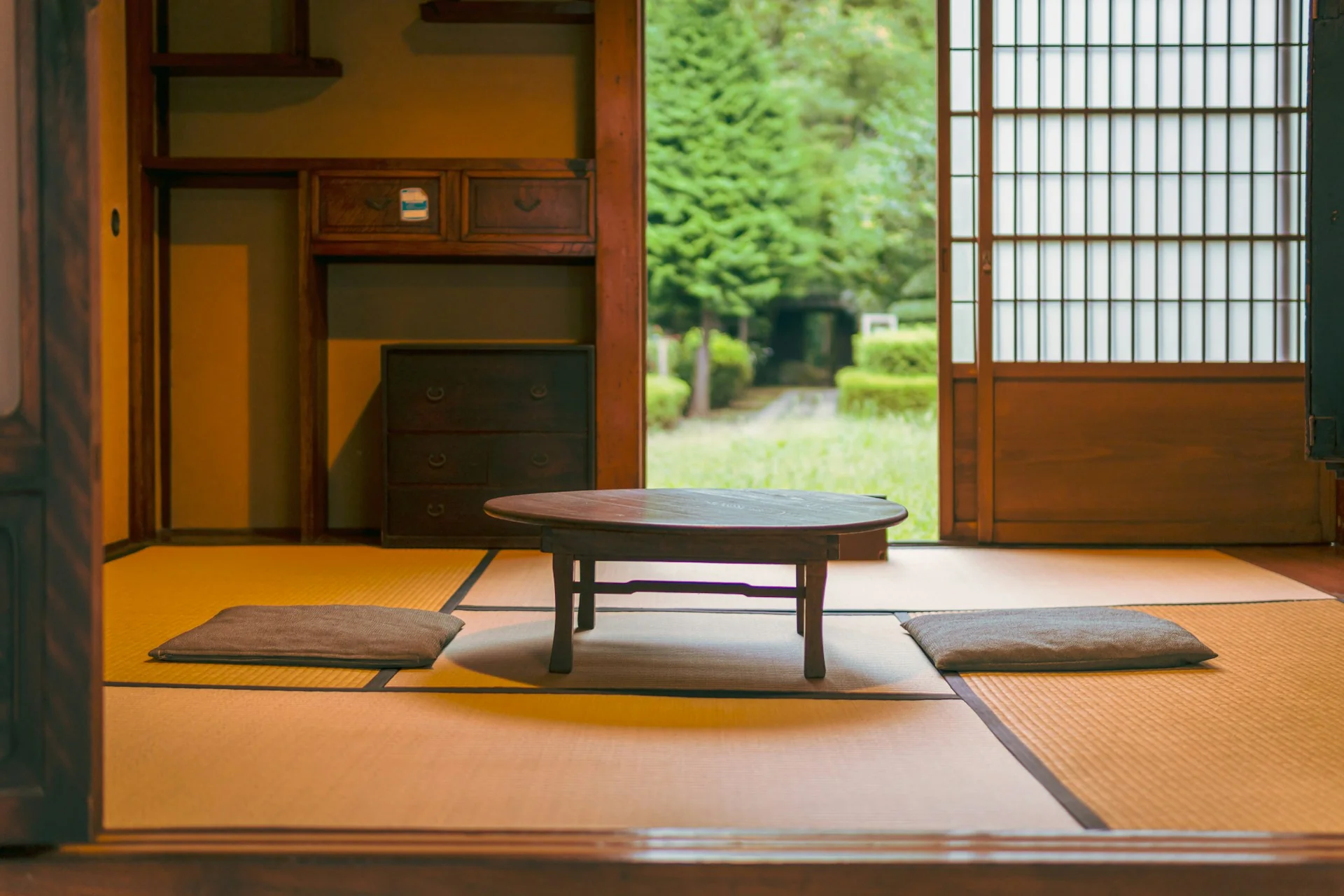 Ruang bertikar tatami dengan meja kayu rendah dan dua bantal duduk di lantai, menghadap pintu geser terbuka ke taman