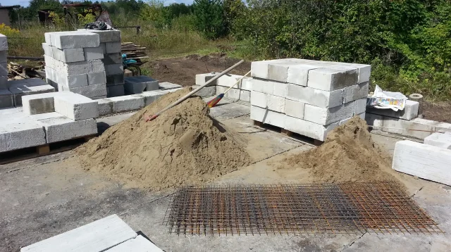 Tumpukan bata ringan AAC putih di atas palet kayu di lokasi proyek, dengan gundukan pasir dan wiremesh di depannya