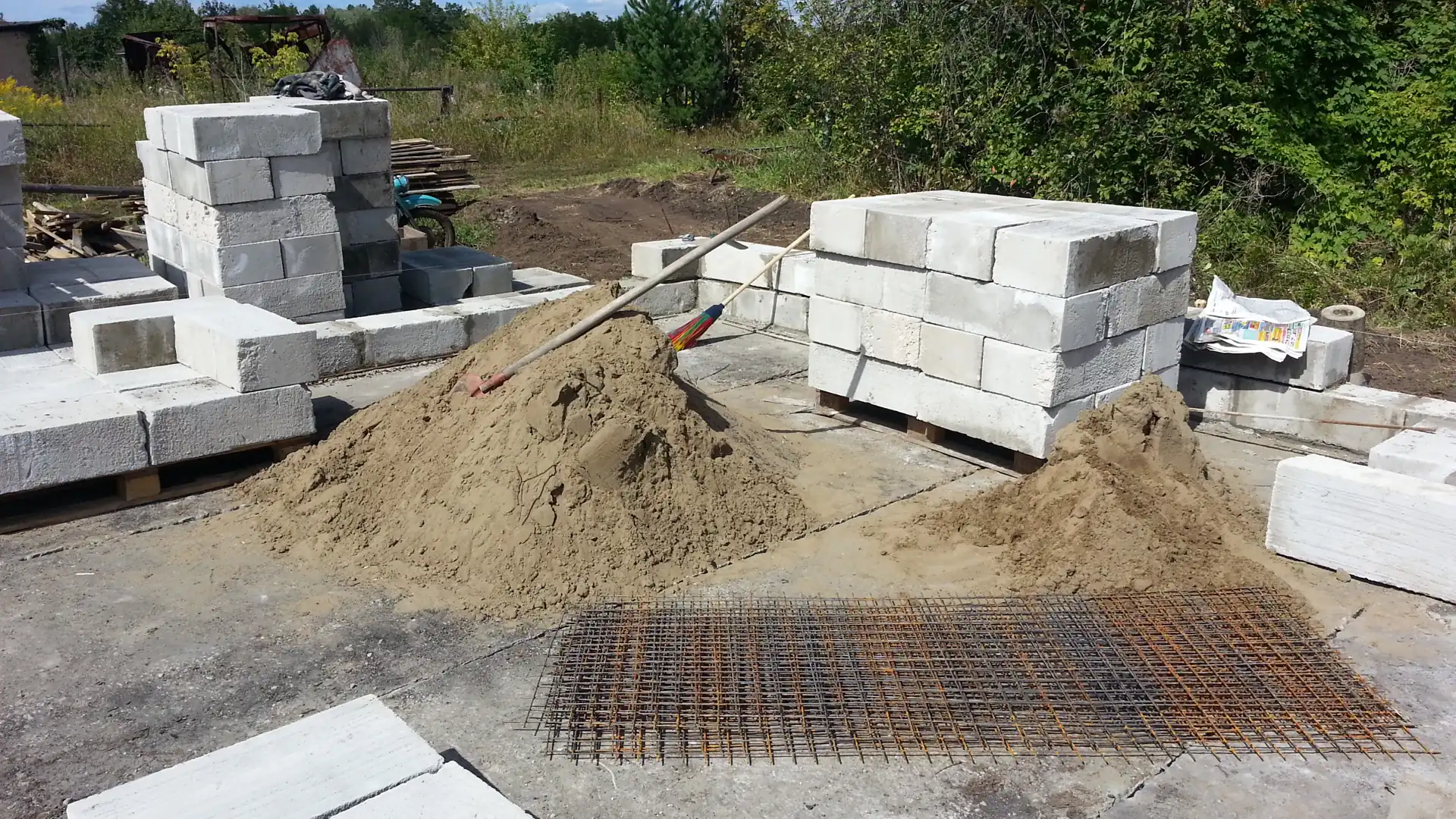 Tumpukan bata ringan AAC putih di atas palet kayu di lokasi proyek, dengan gundukan pasir dan wiremesh di depannya
