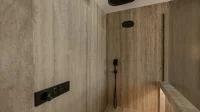 Area shower walk-in dengan dinding keramik besar bermotif batu krem, panel keran hitam, dan bangku berlapis keramik