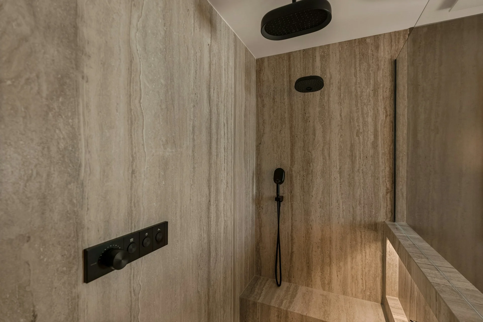 Area shower walk-in dengan dinding keramik besar bermotif batu krem, panel keran hitam, dan bangku berlapis keramik