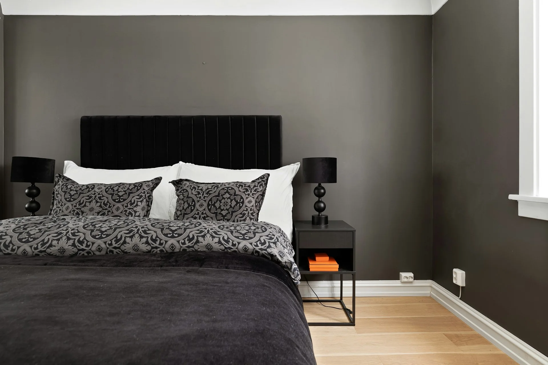 Kamar tidur berdinding abu-abu gelap dengan headboard hitam, selimut gelap, dan lampu meja hitam