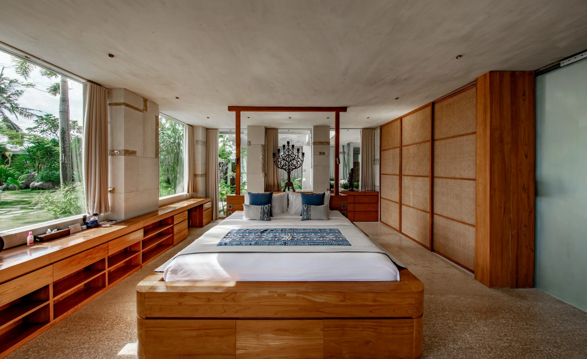 Kamar tidur japandi tropis dengan ranjang platform kayu, linen putih, lemari pintu anyaman rotan, dan jendela lebar ke taman