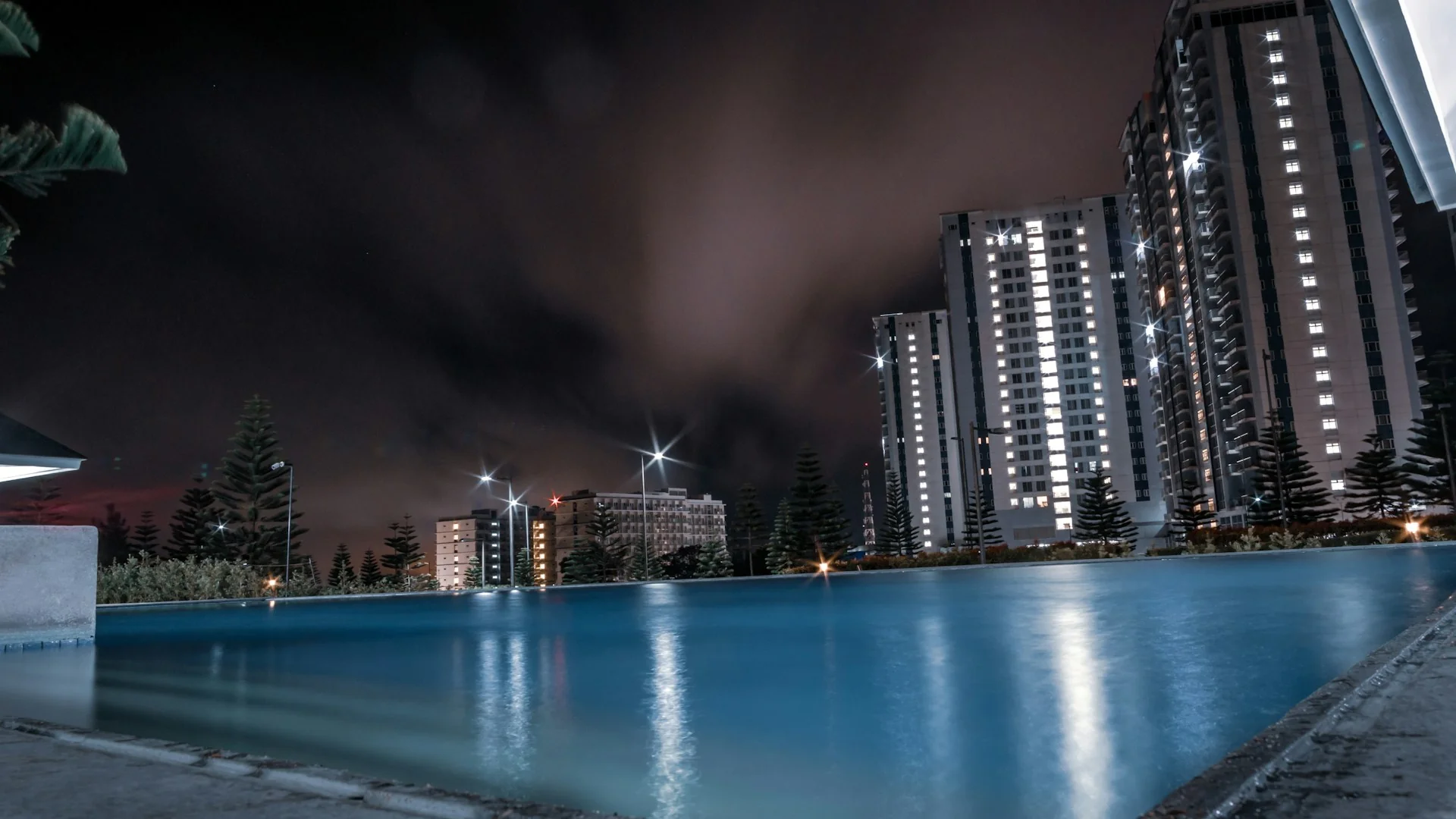 Kolam renang bersama di kompleks apartemen pada malam hari dengan menara hunian di latar belakang