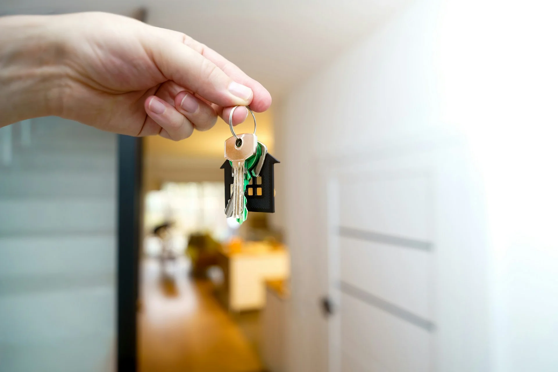 Tangan memegang kunci rumah dengan gantungan berbentuk rumah di depan pintu terbuka