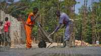 Dua pekerja menuang adukan beton dari gerobak sorong ke lantai cor di antara besi kolom
