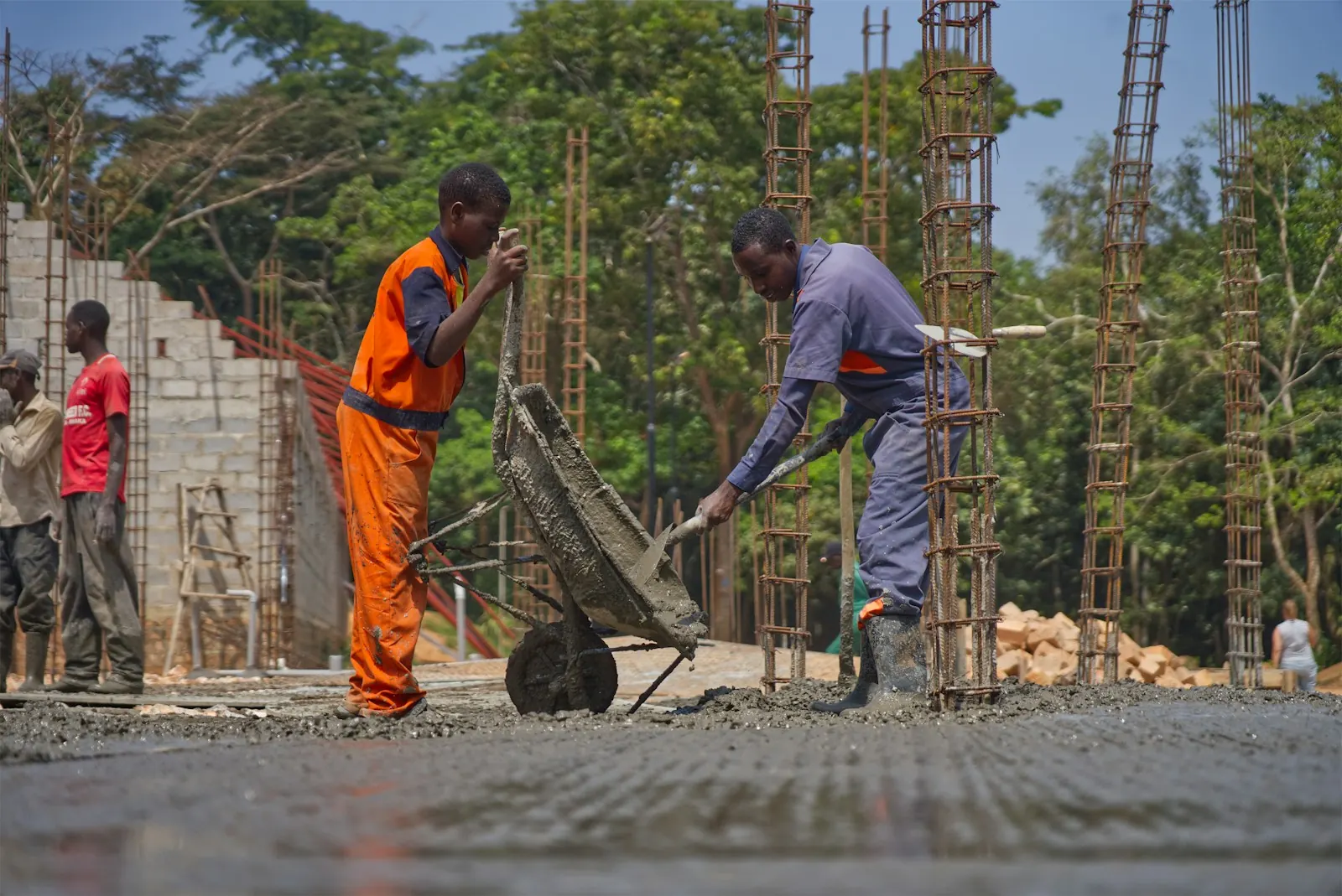 Pekerja menuang adukan beton K-225 Dua pekerja menuang adukan beton dari gerobak sorong ke lantai cor di antara besi kolom