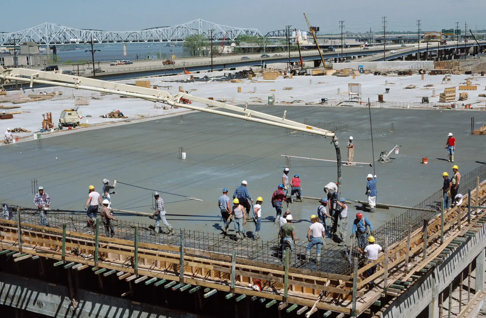 pekerja meratakan beton segar di atas pelat lantai bertulang, dengan pipa pompa beton menuangkan adukan
