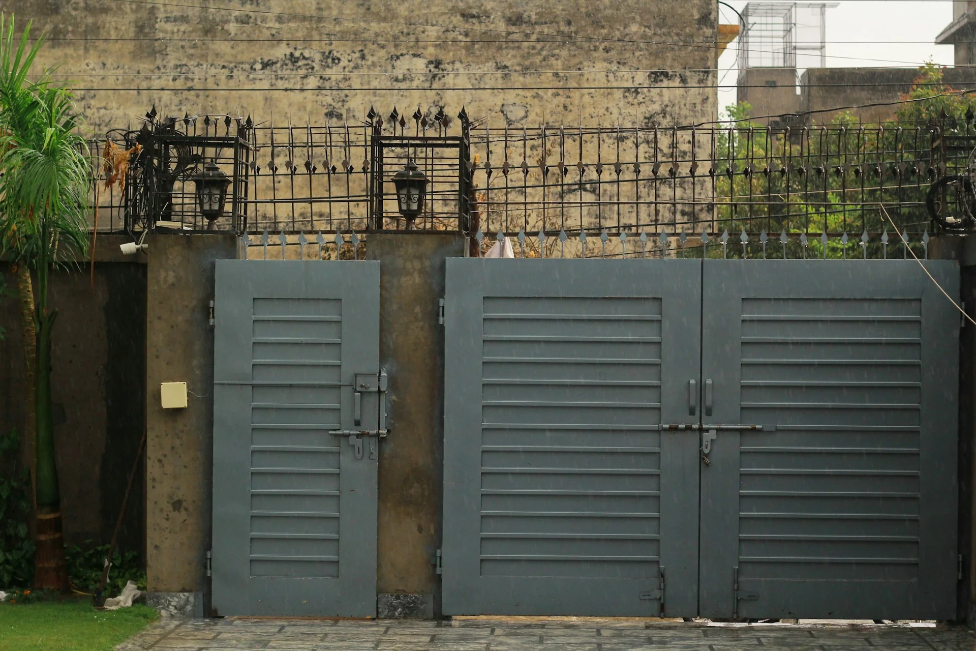 Pintu pagar besi abu-abu dengan bilah horizontal rapat, dua daun besar dan satu pintu kecil di antara pilar
