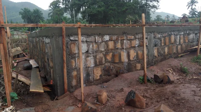 Pondasi batu kali baru selesai dipasang dengan permukaan atas diplester, dikelilingi patok kayu bouwplank dan benang