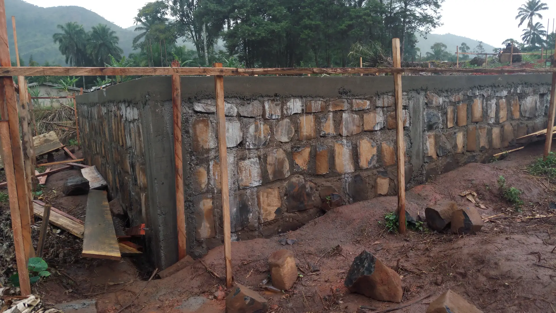 Pondasi batu kali baru selesai dipasang dengan permukaan atas diplester, dikelilingi patok kayu bouwplank dan benang