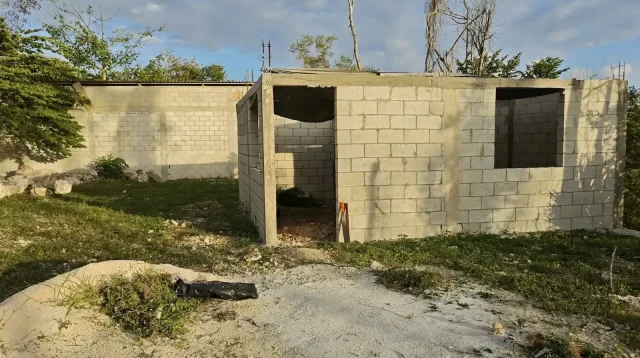Rumah satu lantai berdinding batako yang belum selesai dibangun, tanpa atap, di halaman berumput