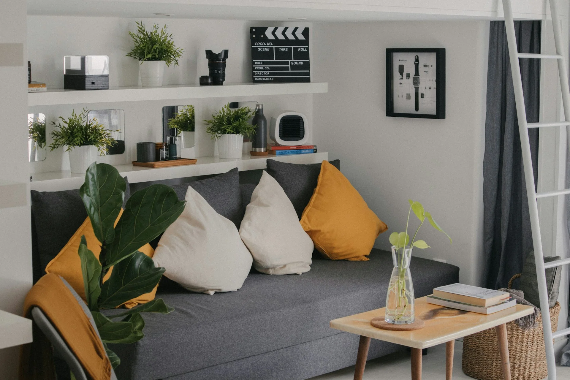 Sofa abu-abu dengan bantal kuning dan putih menempel dinding, dua ambalan putih berisi tanaman dan buku di atasnya, serta meja kayu kecil di depan