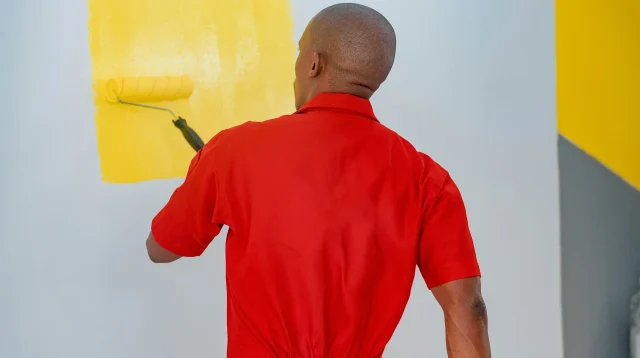 Tukang cat berseragam merah mengecat dinding interior dengan roller warna kuning