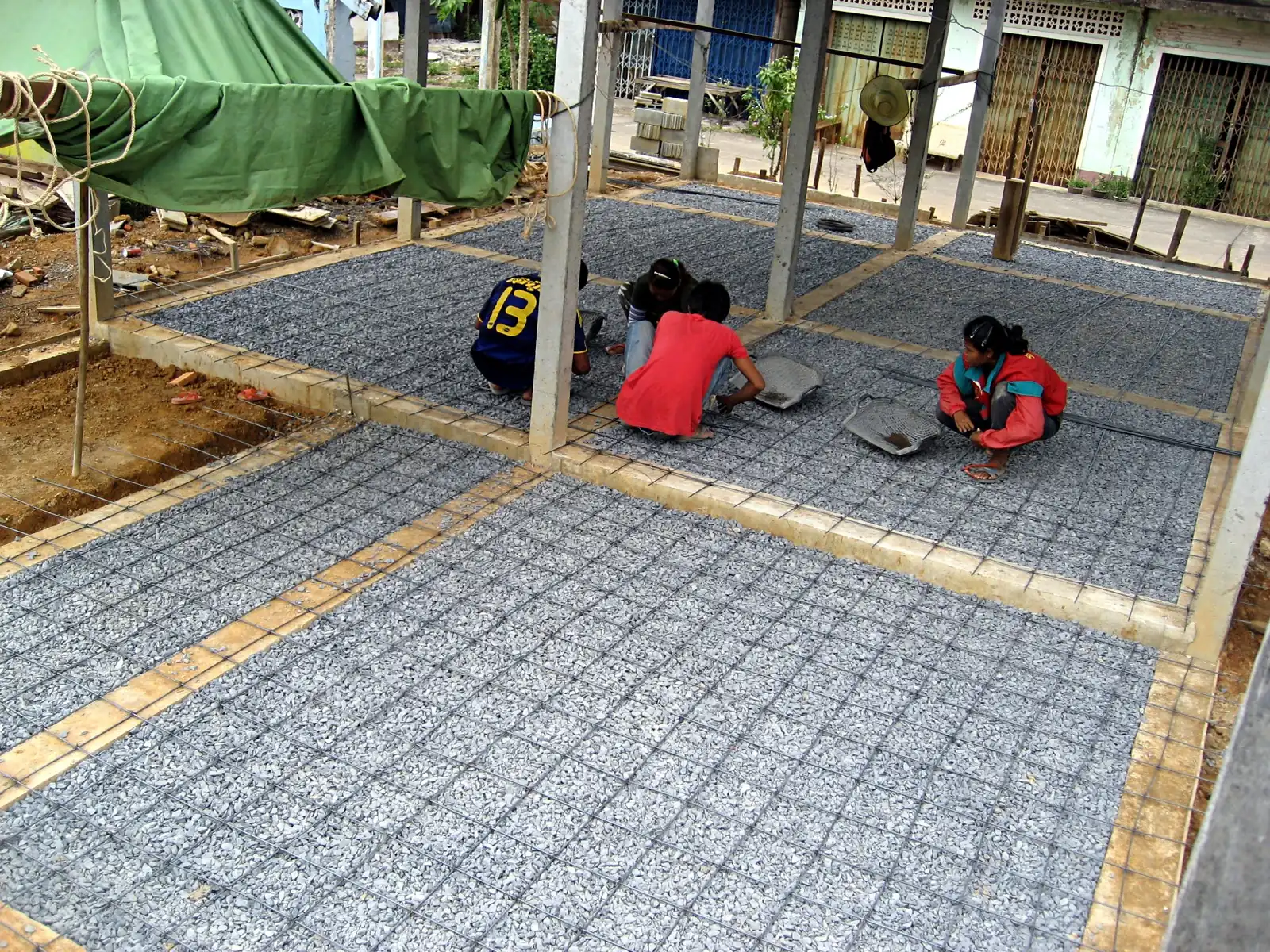 pekerja mengikat jaring besi tulangan pelat lantai di atas lapisan kerikil sebelum dicor, di antara kolom beton rumah