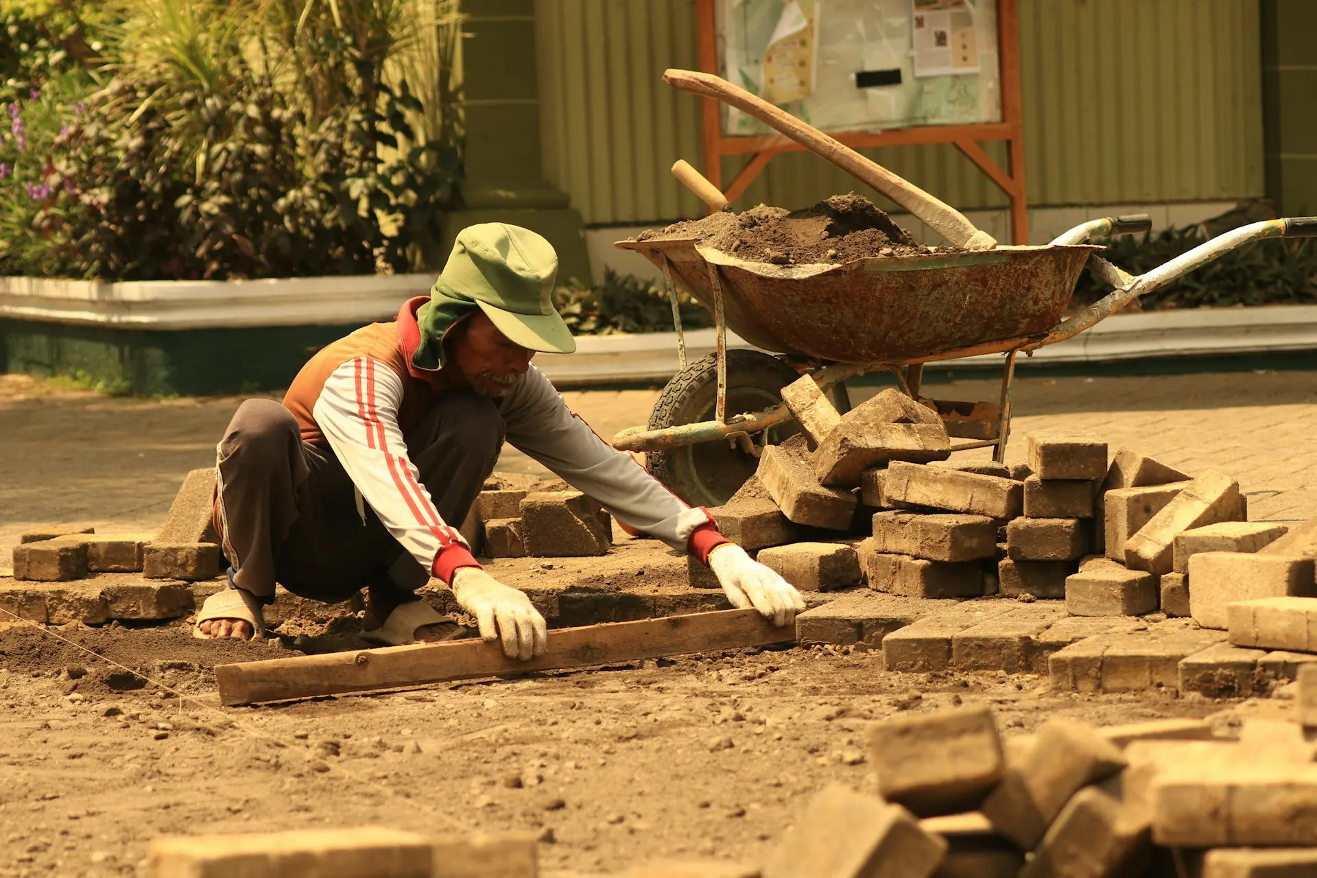 upah-tukang-jabodetabek-pasang-paving upah tukang jabodetabek: tukang berjongkok memasang bata paving dengan papan perata di samping gerobak dorong